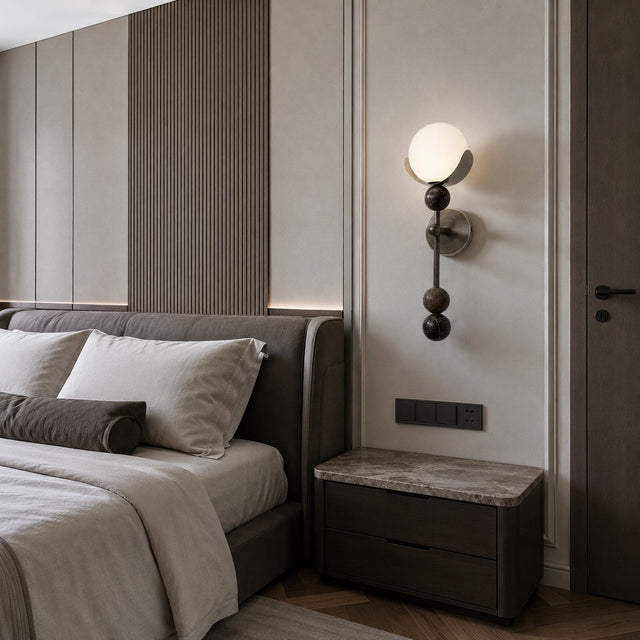 A modern bedroom interior featuring a bed with neutral-toned bedding, a dark wood nightstand with a stone-look top, a wall-mounted spherical sconce, and a paneled wall background.