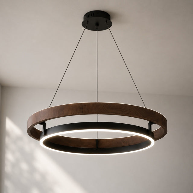 A modern two-ring circular pendant light with a dark wood outer ring and a black inner ring, suspended by thin cables from a black ceiling mount against a light gray wall.
