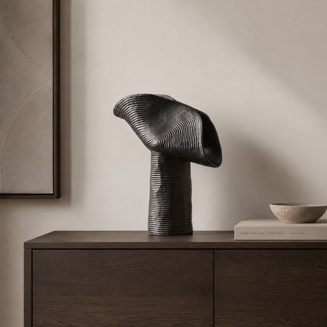A sculptural black ceramic table lamp with a ribbed cylindrical base and a flowing, open flame-like top, displayed on a dark wood sideboard against a warm beige wall.