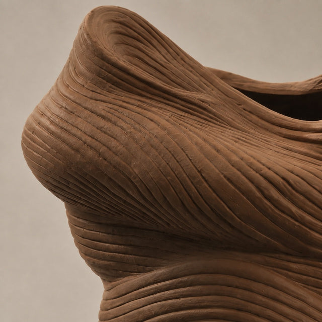 A close-up of a sculptural brown textured object with flowing, ribbed contours against a plain light beige background.