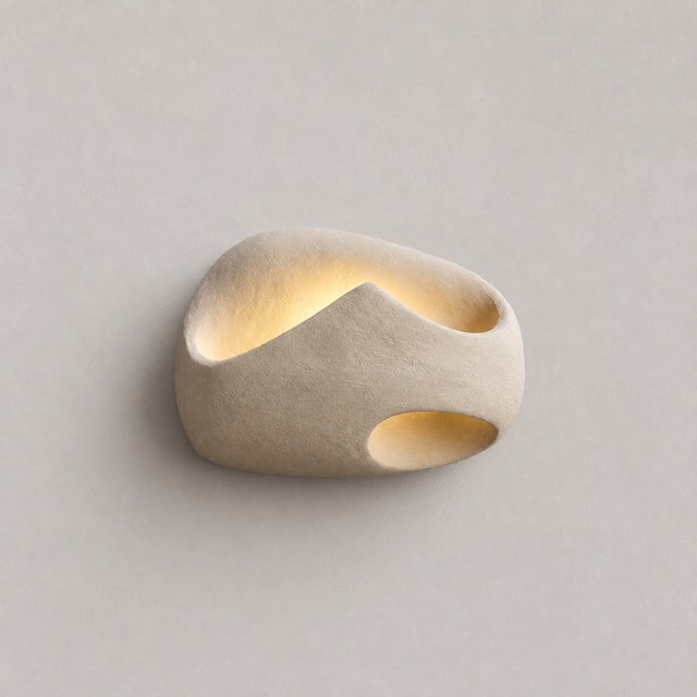 A modern, organic-shaped wall sconce in a textured beige stone or concrete finish, featuring integrated warm white LED lighting in its recessed channels, set against a solid light gray background.