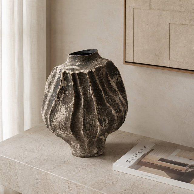 A textured, ribbed ceramic vase in a dark charcoal gray with lighter beige highlights, displayed on a light stone console table next to a magazine on a beige wall.